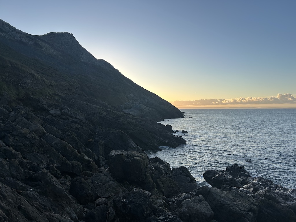 Rocky cove at sunrise. The sun is behind the cliff so the rocks are dark but the sky is blue and there is a orange glow on the horizon. The tide is high.