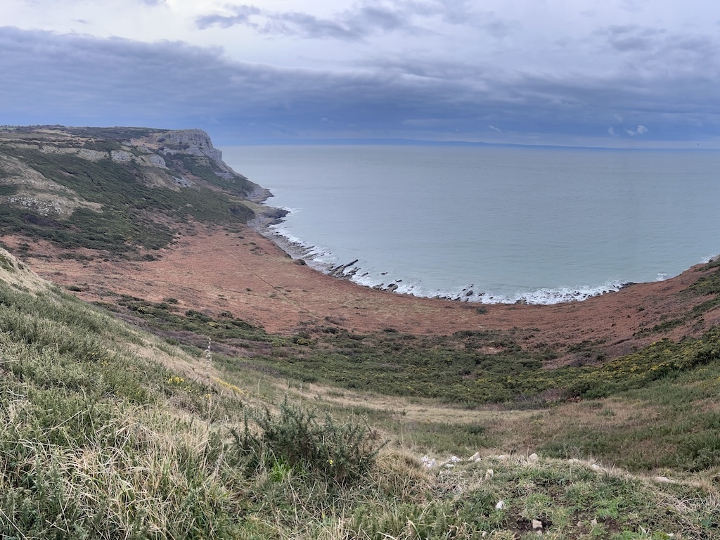 Looking down a wide sweeping valley to a rocky bay. The sky is grey but there are nice muted tones of red, green and blue.